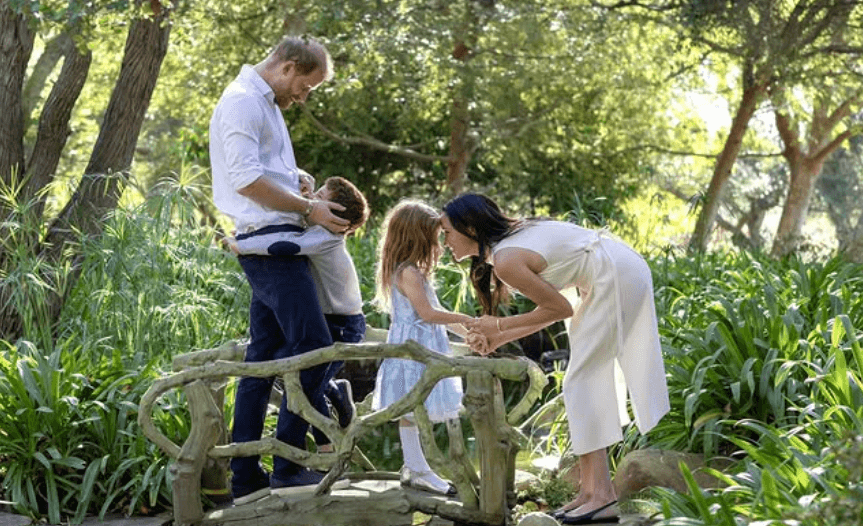 Meghan Markle and Prince Harry sit together on a picnic blanket with Lilibet, surrounded by greenery and flowers.