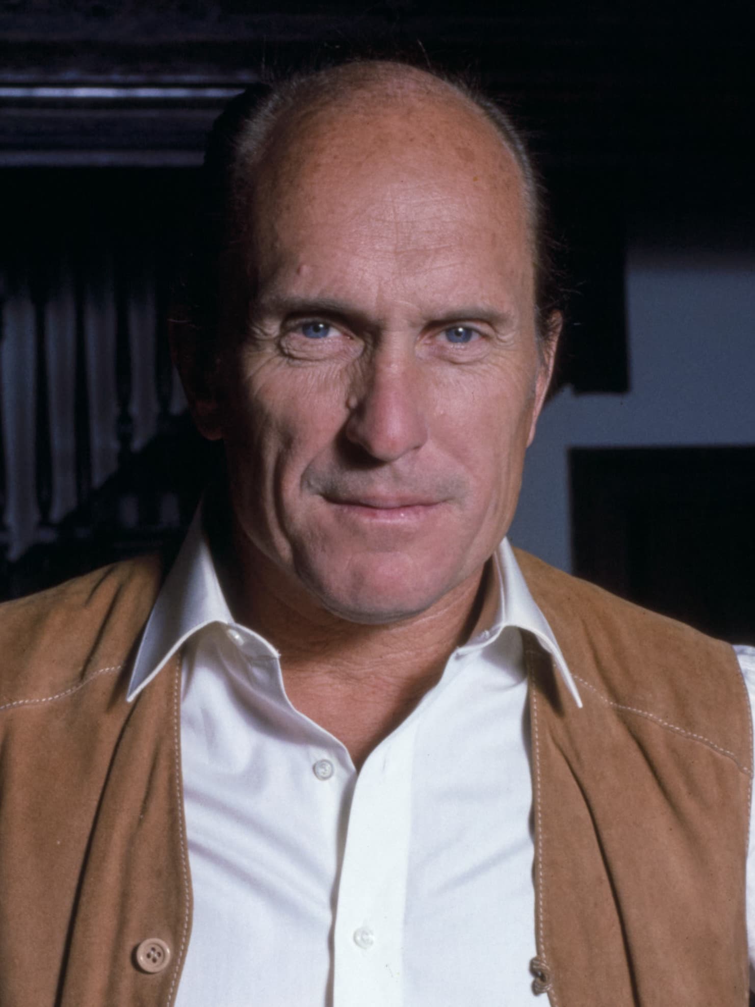 Robert Duvall smiling while wearing a black suit and tie at a red carpet event with a vintage film backdrop.