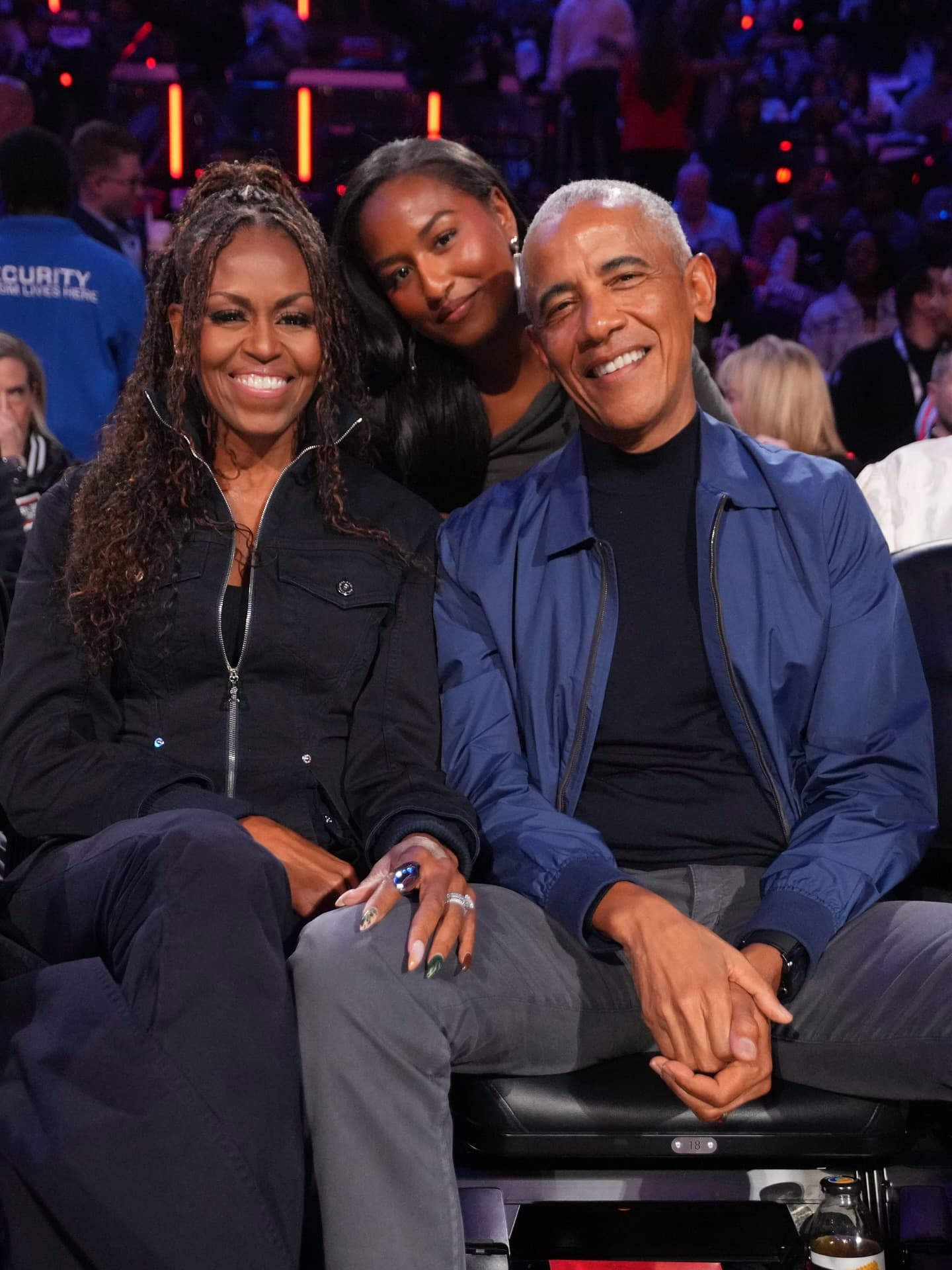 The Obamas sitting together in a crowd at the NBA All-Star Weekend, dressed casually in jackets and jeans, smiling and enjoyi