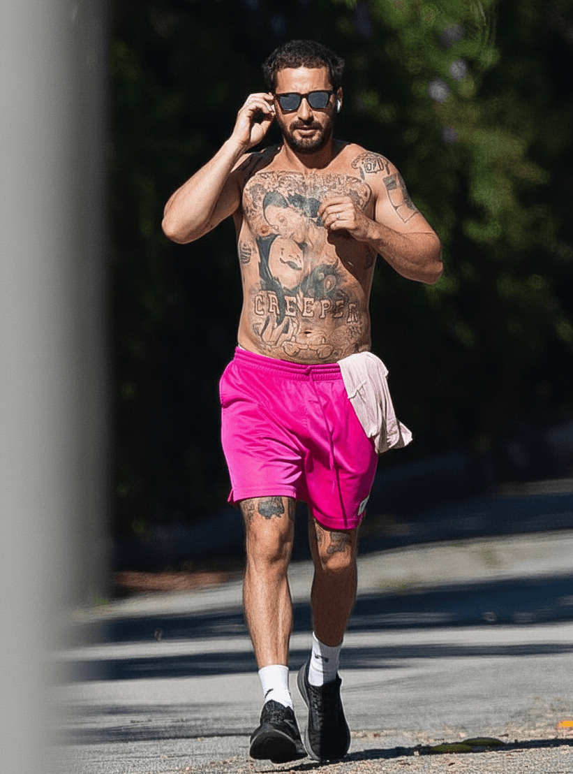 Shia LaBeouf walking alone down a city street, wearing a black hoodie and distressed jeans, looking contemplative.