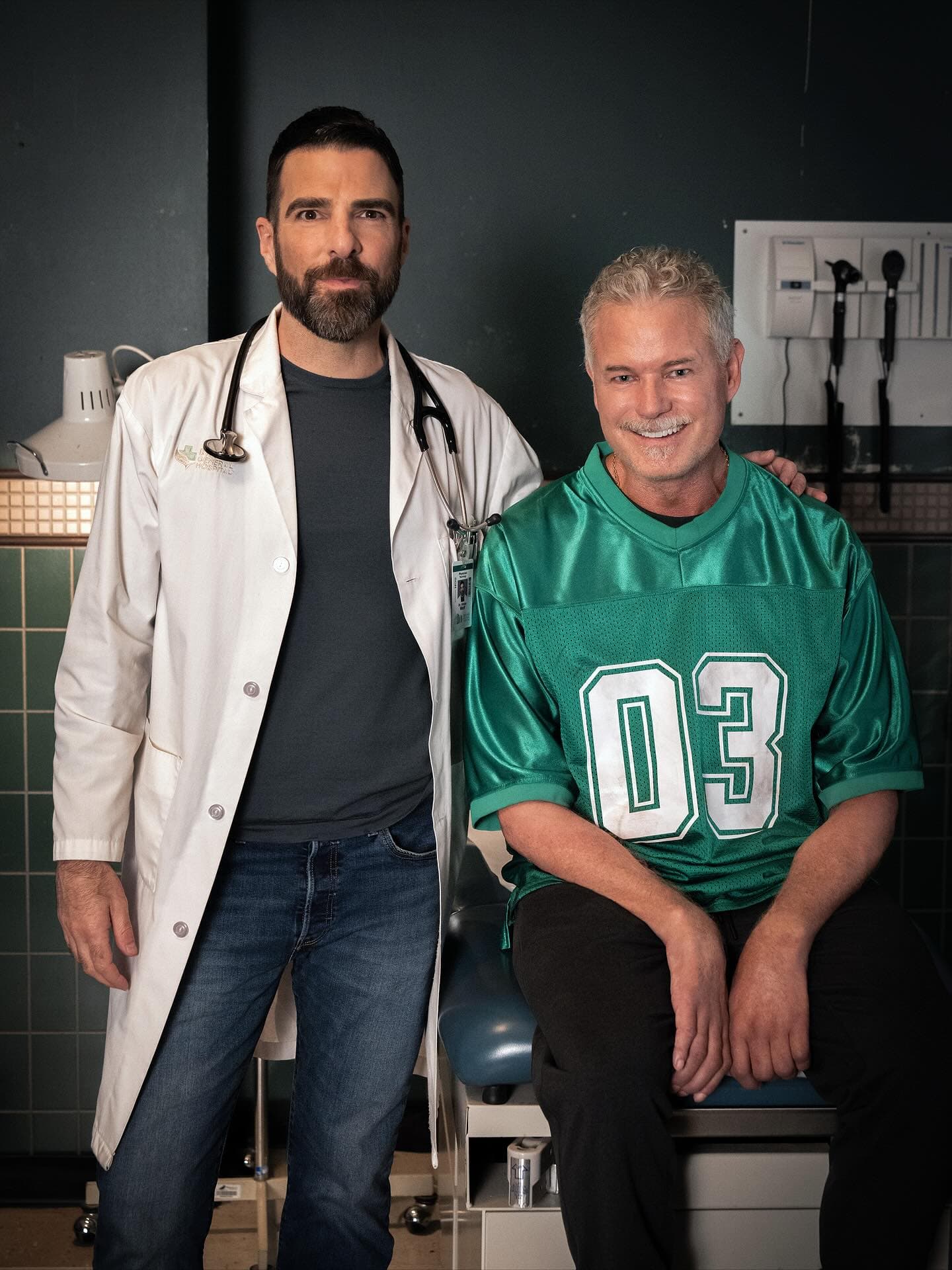 Eric Dane in a black suit, standing on stage with a solemn expression, holding a microphone during a tribute event.