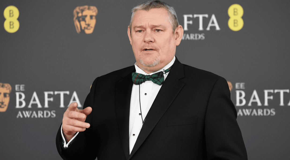 John Davidson wearing a tuxedo on stage at the BAFTAs, gesturing with hands and speaking into a microphone.