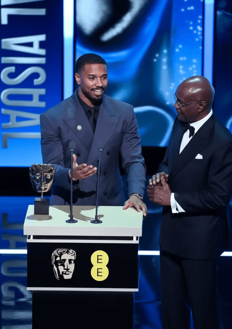 John Davidson speaking at the BAFTAs wearing a tailored black suit and white shirt, gesturing animatedly on stage.