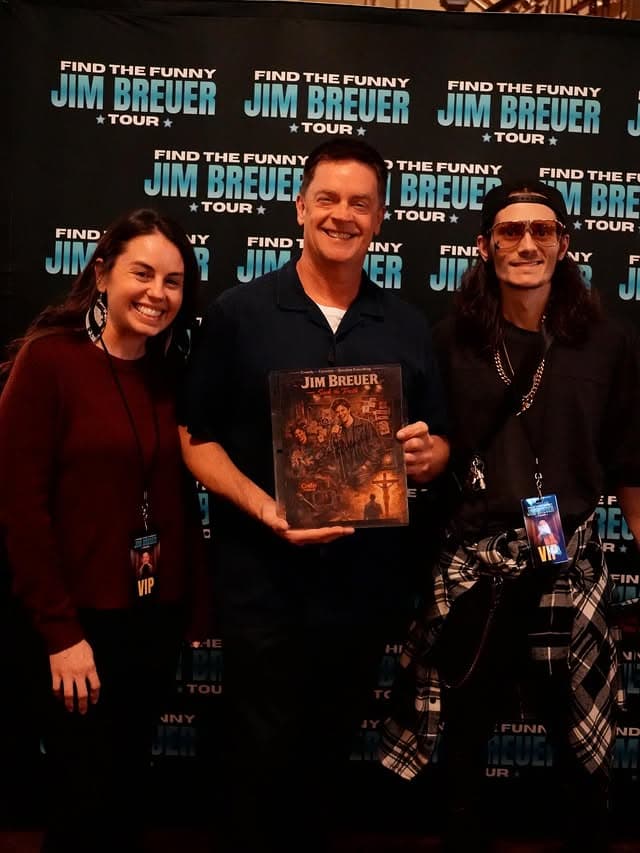 Jim Breuer sitting in an airport lounge wearing a casual t-shirt and jeans, looking frustrated while holding a coffee cup.