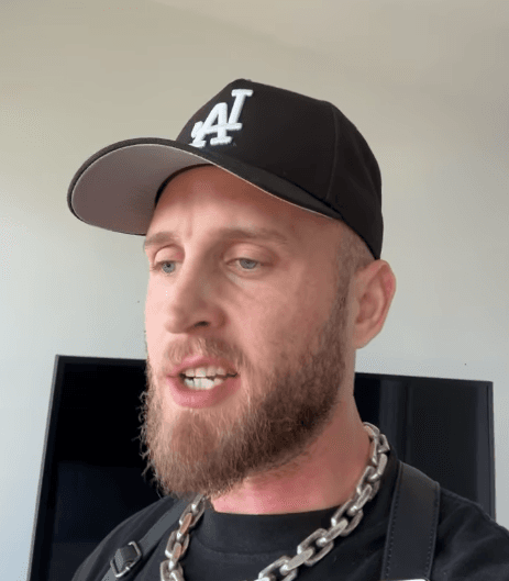 Chet Hanks sitting on a suitcase at a busy airport terminal, wearing a casual white t-shirt and black pants.