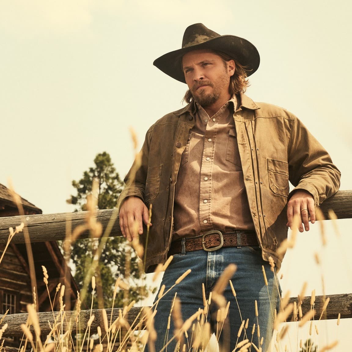 A cowboy in a wide-brimmed hat stands beside a horse in a dusty landscape during sunset, with mountains in the background.