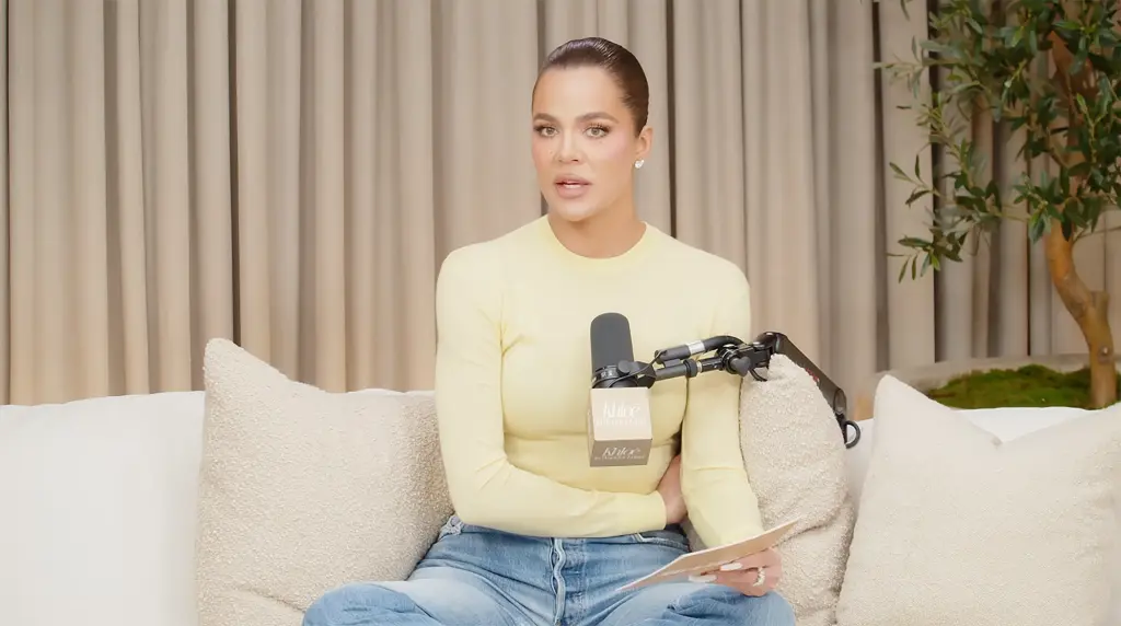 Khloé Kardashian sitting on a couch, wearing a white sweater, with a thoughtful expression and a coffee cup in her hand.