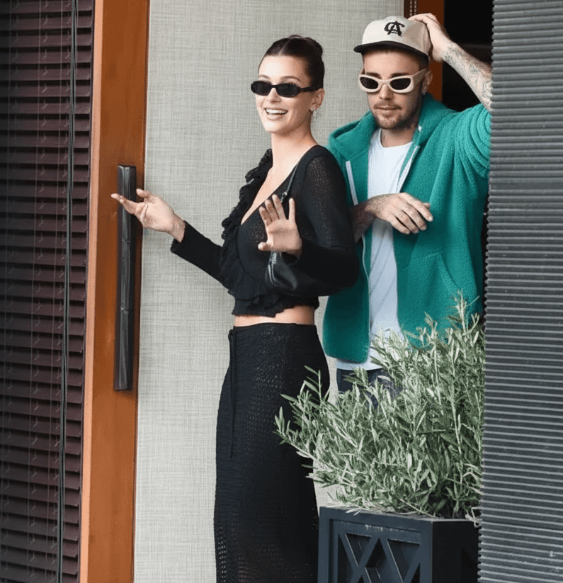 Justin and Hailey Bieber smiling and holding hands, both wearing casual outfits, in a sunlit outdoor setting.