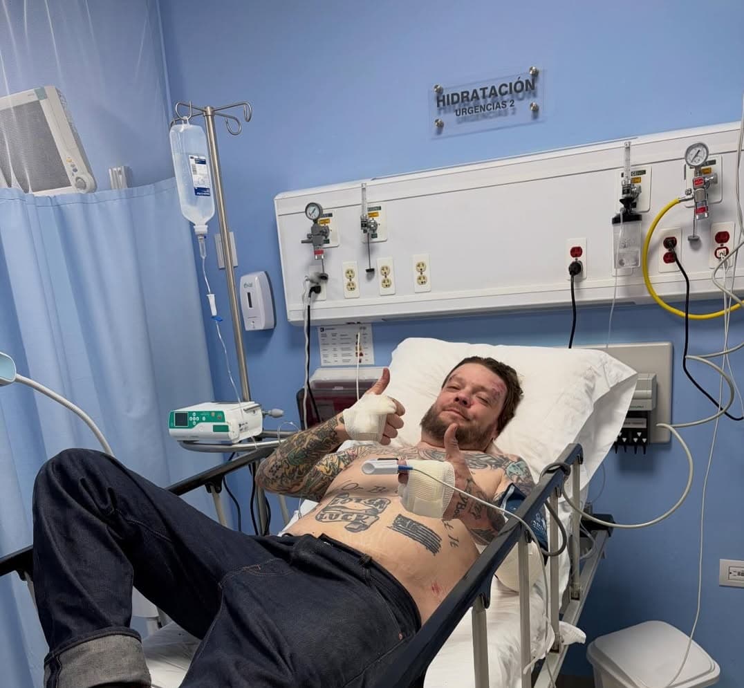 Rick Harrison standing in a brightly lit shop, arms crossed, wearing a black shirt and jeans, looking intently at the camera.