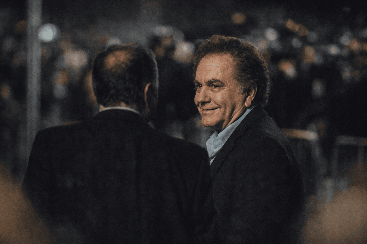 A group of elegantly dressed Hollywood celebrities at a lavish event, chatting and laughing under sparkling chandeliers.